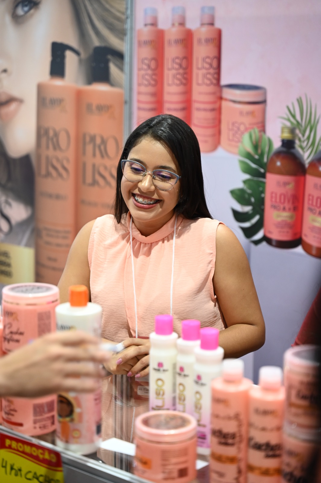 Jovem de óculos e blusa salmão sorri atrás de um balcão com produtos capilares em tons de rosa
