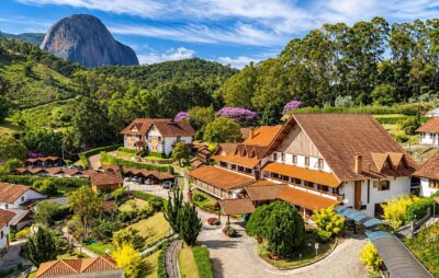 Vista de hotel e chalés com telhados laranja em meio a jardins e mata, com uma grande formação rochosa ao fundo sob céu azul