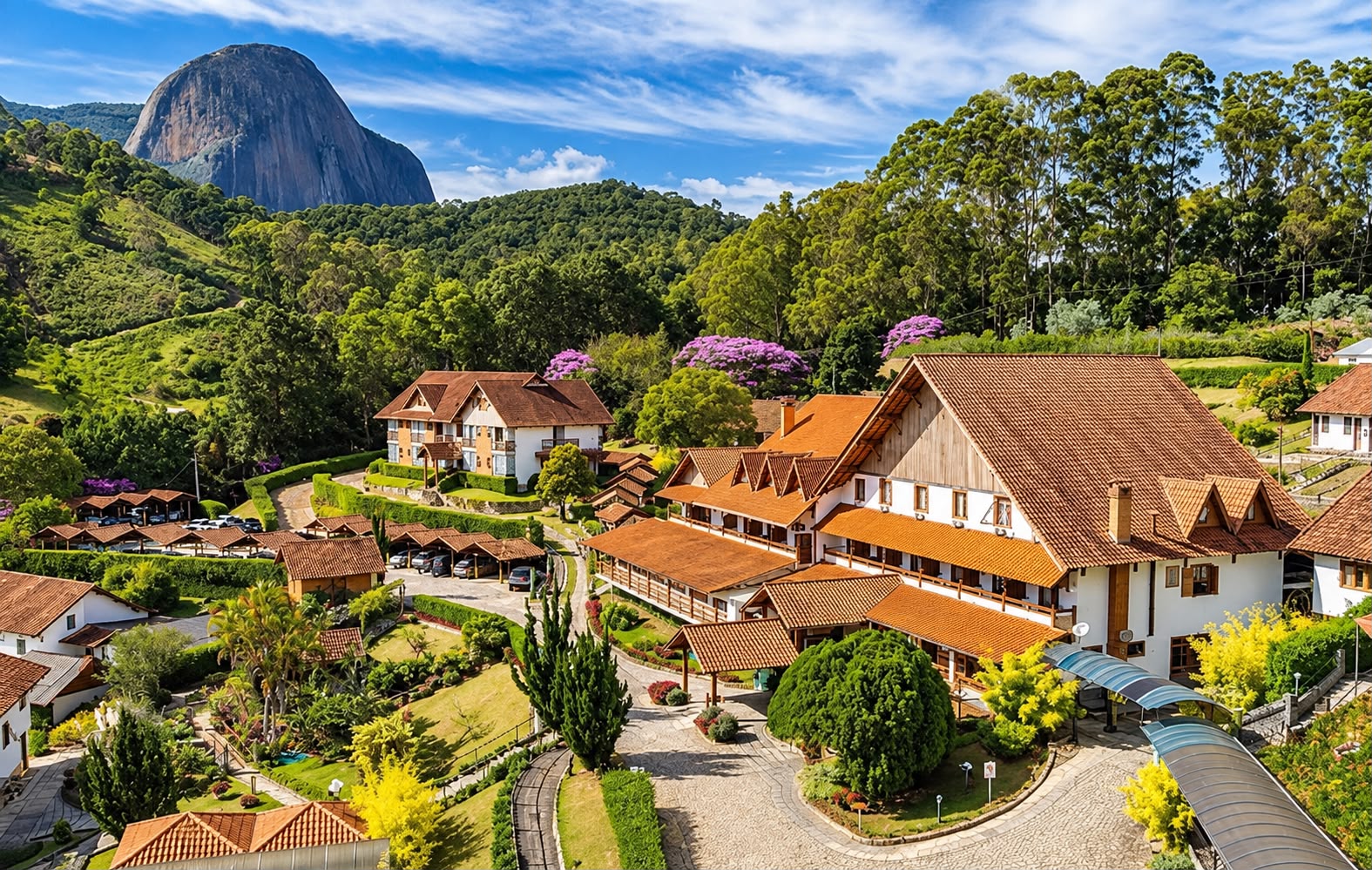 Vista de hotel e chalés com telhados laranja em meio a jardins e mata, com uma grande formação rochosa ao fundo sob céu azul