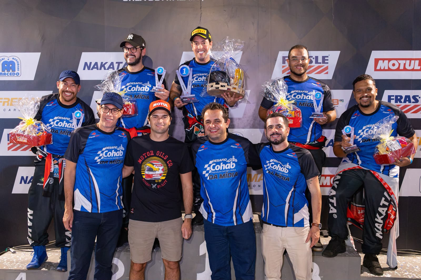 Participantes do Desafio Cohab Premium da Mídia com camisas azuis do evento, troféus e cestas de brindes no pódio do Piquet Kart, ao lado do piloto Nelsinho Piquet, de camiseta preta