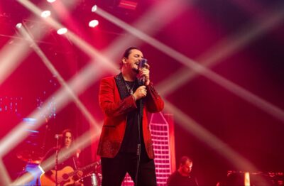 O cantor Dino Fonseca, de paletó vermelho brilhante, canta em um microfone retrô no palco, com luzes vermelhas e um violonista ao fundo