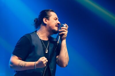 O cantor Dino Fonseca, de colete escuro e camiseta preta, canta de olhos fechados segurando um microfone retrô, sob luz azul no palco