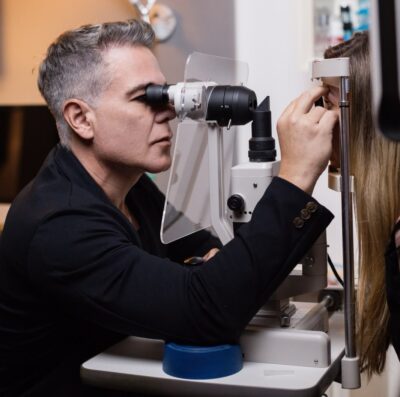 Oftalmologista de cabelos grisalhos e blazer preto examina os olhos de uma paciente com um equipamento de lâmpada de fenda