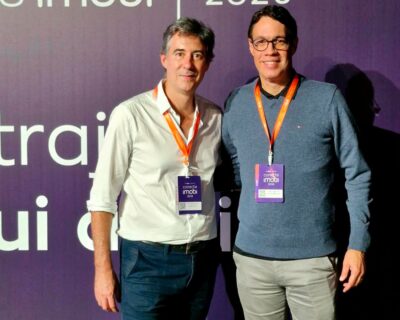 Dois homens com crachás do Conecta Imobi 2026 posam lado a lado diante de um painel roxo do evento