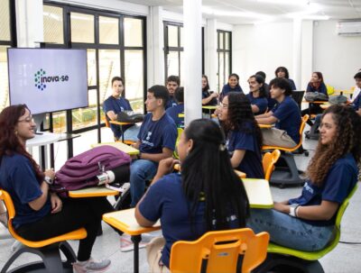 Jovens com camisetas azul-marinho sentados em cadeiras laranja e verdes em sala de aula, com uma TV exibindo a logo do Inova-SE
