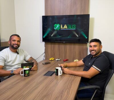 Dois homens sorriem sentados a uma mesa de madeira com canecas do L.A Talks, diante de uma TV com a logomarca do podcast