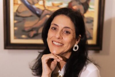 A advogada Luceli Lemos Botezel, de cabelos escuros e blusa branca, sorri apoiando o queixo na mão, diante de um quadro colorido