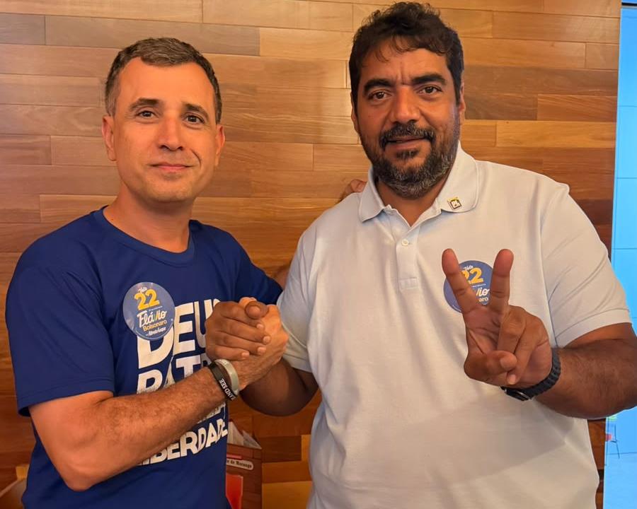 Dois homens apertam as mãos e posam lado a lado em frente a uma parede de madeira, um de camiseta azul e outro de camisa polo branca fazendo sinal de V, ambos com adesivo de campanha do 22