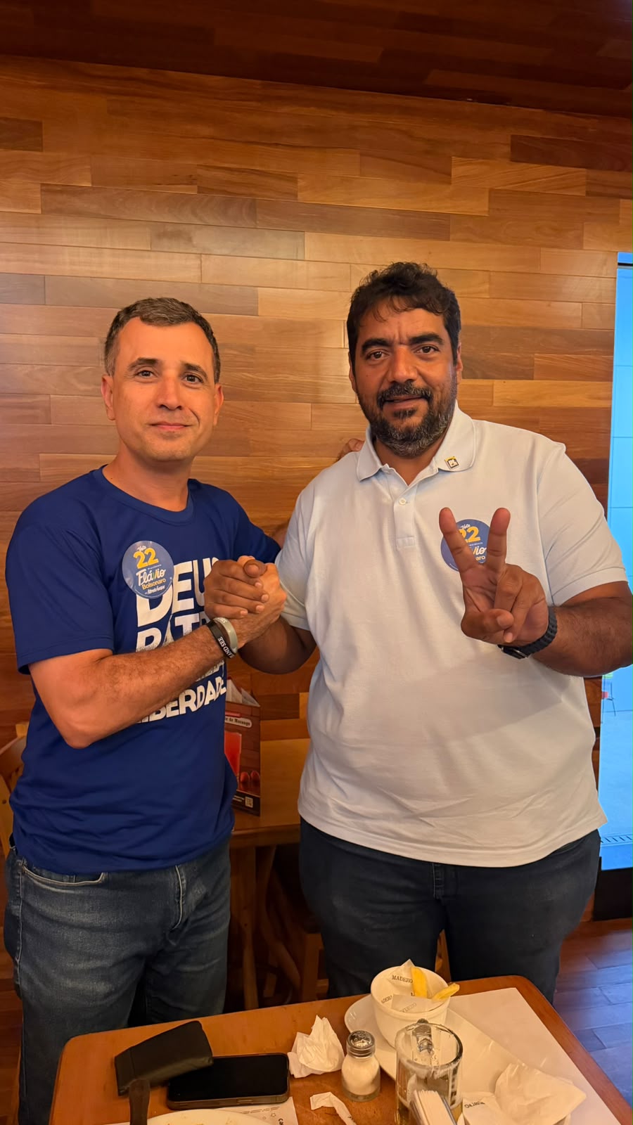 Foto vertical de dois homens de pé apertando as mãos ao lado de uma mesa de restaurante, com parede de madeira ao fundo