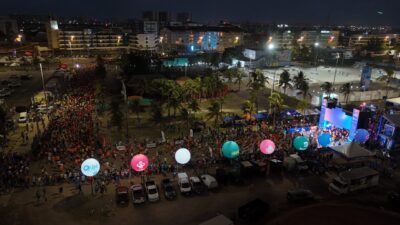 Vista aérea noturna de milhares de corredores com camisetas laranja e azuis em uma avenida de Aracaju, com palco iluminado, balões e prédios ao fundo