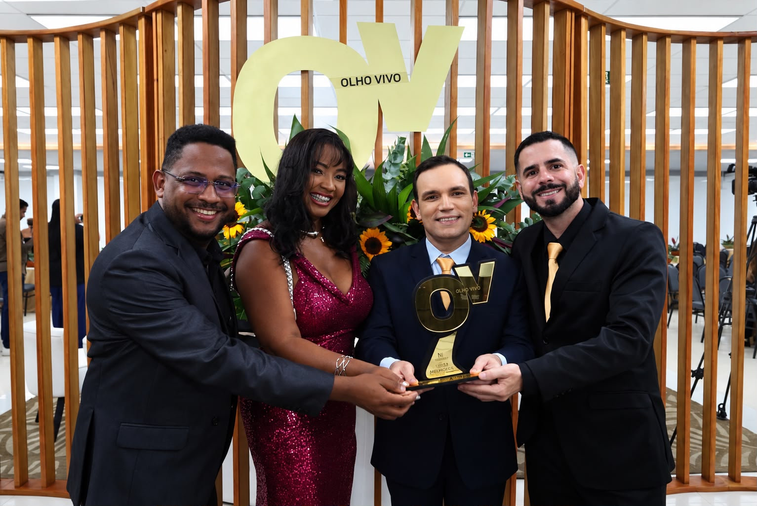 Quatro pessoas em trajes formais sorriem segurando juntas o troféu dourado do Prêmio Olho Vivo, diante de um painel de madeira com a logo OV