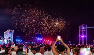 Multidão filma com celulares a queima de fogos dourados no céu noturno durante festa de Réveillon, com palco e torres de luz ao fundo
