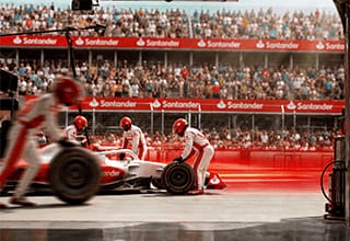 Equipe de mecânicos de macacão branco e vermelho faz pit stop em carro de corrida, com arquibancada cheia e placas do Santander ao fundo