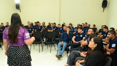 Mulher com camiseta lilás da SerMulher fala para um grupo de guardas municipais de farda azul sentados em uma sala