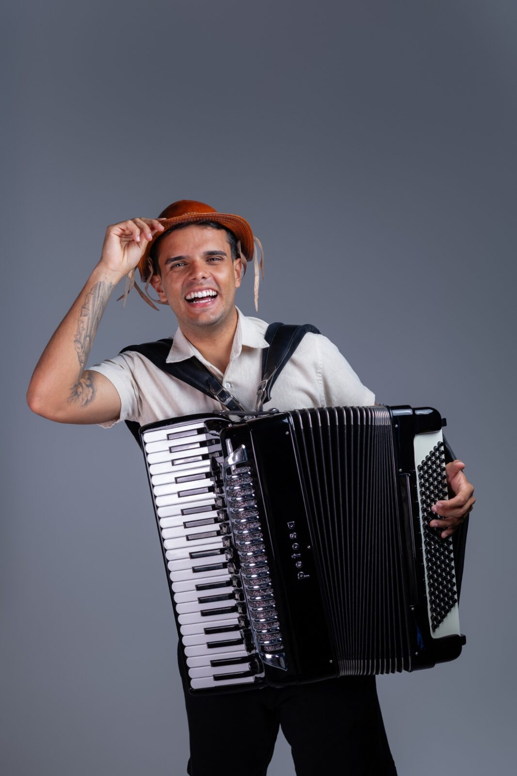 O cantor William Sanfona sorri segurando um chapéu de couro com uma das mãos e uma sanfona preta na frente do corpo, em fundo cinza