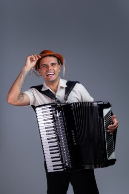 O cantor William Sanfona sorri segurando um chapéu de couro com uma das mãos e uma sanfona preta na frente do corpo, em fundo cinza