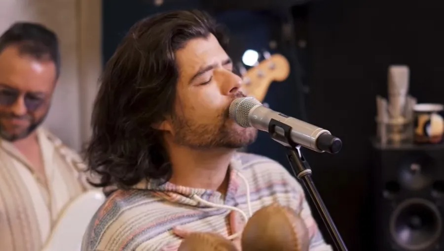 Cantor de cabelos longos canta de olhos fechados em microfone dentro de um estúdio, com outro músico ao fundo